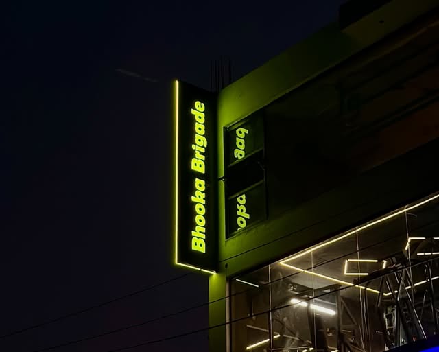 Vertical illuminated Bhooka Brigade sign on the corner of a shop at night.