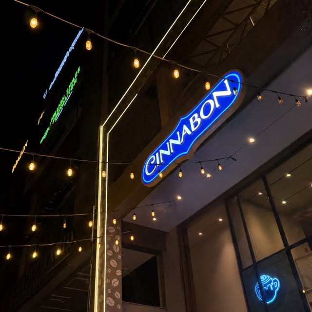 Blue illuminated Cinnabon blade sign at night, with a small neon cup in the window.